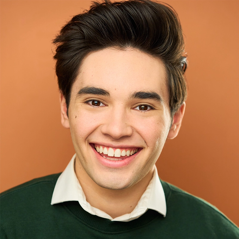 A young person with short, dark hair smiles widely, showing teeth. They wear a dark green sweater over a white collared shirt. The background is a warm, solid light brown. The lighting is bright, highlighting their cheerful expression.