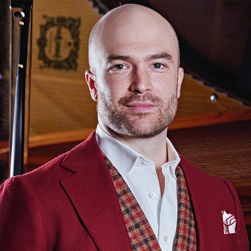 A bald man with a trimmed beard is wearing a white dress shirt, a patterned vest, and a red suit jacket with a pocket square. He is standing indoors in front of a stringed musical instrument, likely a grand piano. The lighting is warm and soft.