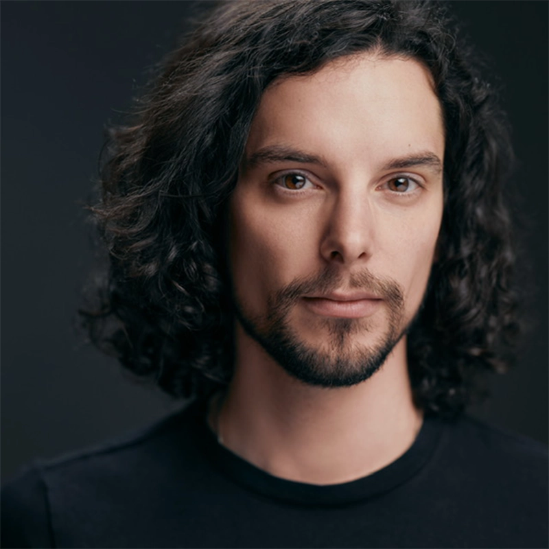 A person with shoulder-length curly dark hair and a short beard looks directly at the camera. They have light skin and are wearing a plain black shirt. The background is blurred and dark, emphasizing their face. The lighting is soft and even.