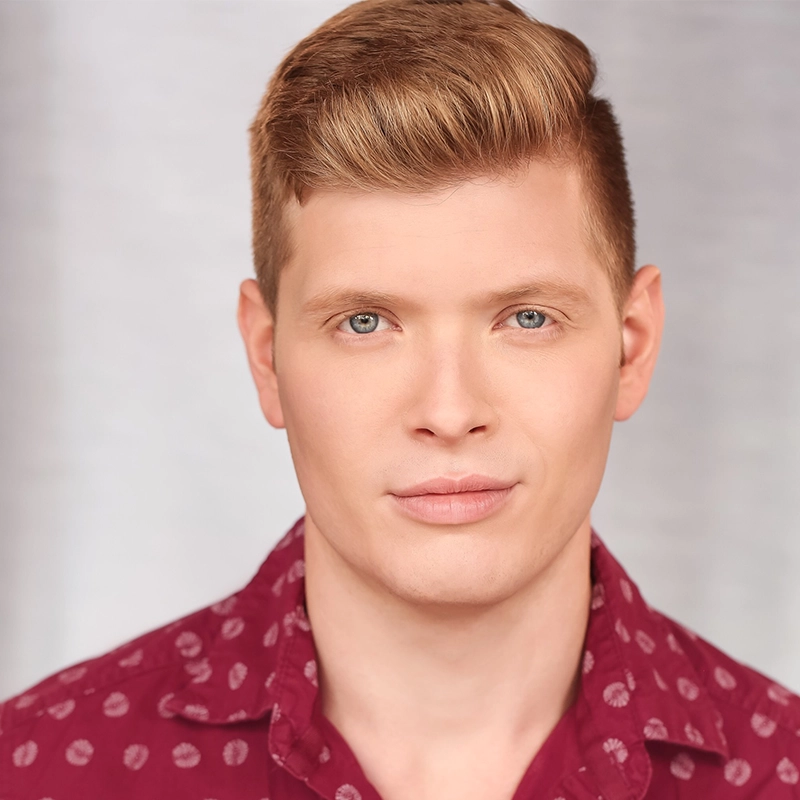 A young man with fair skin and short, neatly styled light brown hair stares into the camera with a neutral expression. He wears a maroon button-up shirt with a white polka dot pattern. The softly blurred background evokes Katie Yeomans’ subtle style.