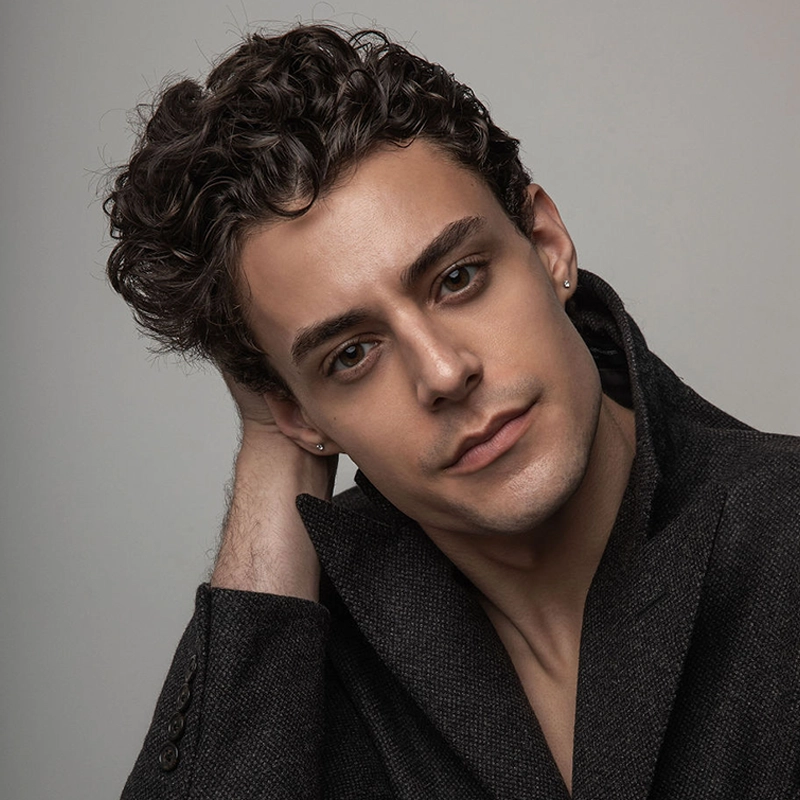 A young man with short, curly dark hair and light skin gazes at the camera, head tilted to one side and hand behind his neck. Julian Manjerico wears a dark textured coat with a wide collar and stud earrings, set against a plain neutral background.