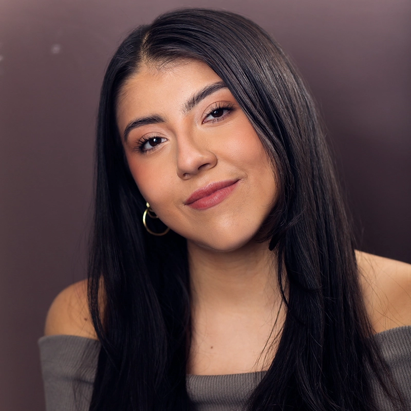 Aidan Michael Kelly captures a young woman with long straight black hair, smiling softly at the camera. She wears gold hoop earrings and an off-the-shoulder gray top, framed by a plain, muted brown background.