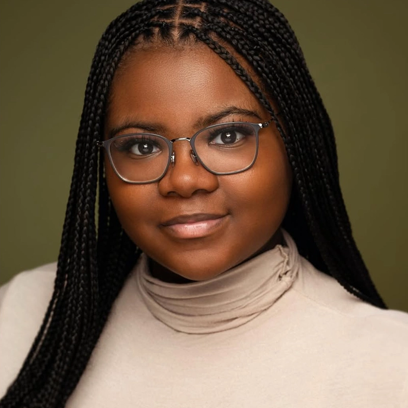 Vivica Powell, a young woman with dark braided hair and glasses, smiles softly in a beige turtleneck. Her smooth skin and dark eyes stand out against the plain olive green background in this well-lit, professional portrait.