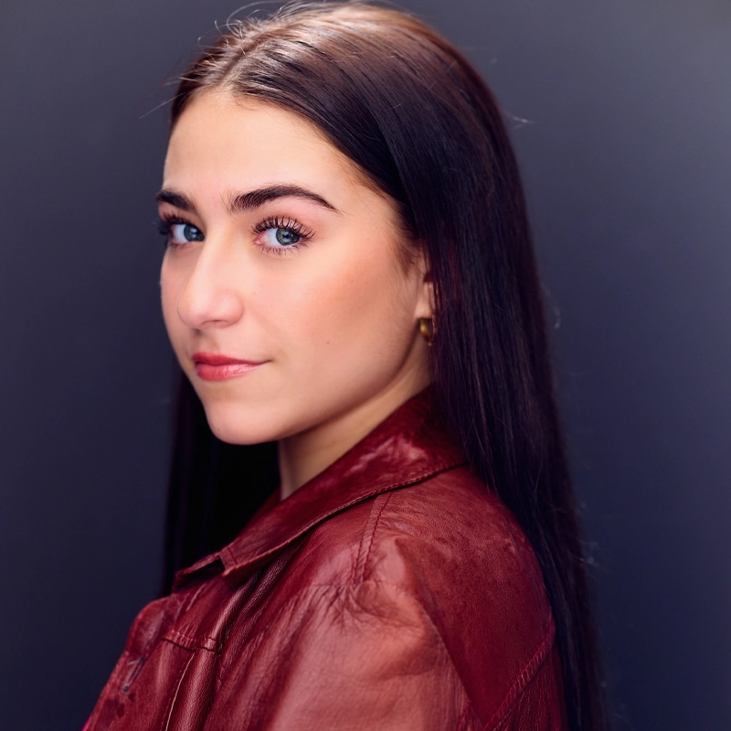 Lyla Karekinian, a young woman with long straight brown hair and blue eyes, looks over her shoulder towards the camera. She wears a red leather jacket and small gold hoop earrings, softly lit against a plain, dark background to highlight her face.