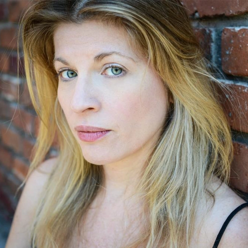 A woman with long blonde hair and light green eyes stands against a red brick wall, looking directly at the camera with a neutral expression. She wears a black top with thin straps and natural makeup. The lighting is soft and highlights her facial features.
