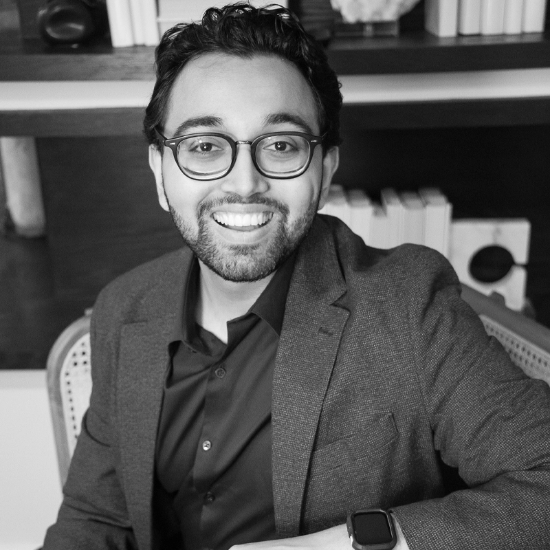 A smiling man with short dark hair, a beard, and glasses sits on a cane-backed chair. He wears a dark blazer, shirt, and a smartwatch. Books and decorative items are on shelves behind him. The photo is black and white.