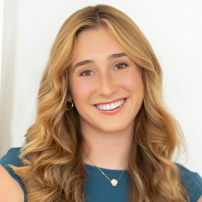 A young woman with long, wavy blond hair smiles at the camera. She wears a teal top, gold hoop earring, and a necklace with a round pendant—her serene style reminiscent of an Aidan Michael Kelly portrait against a plain white background.