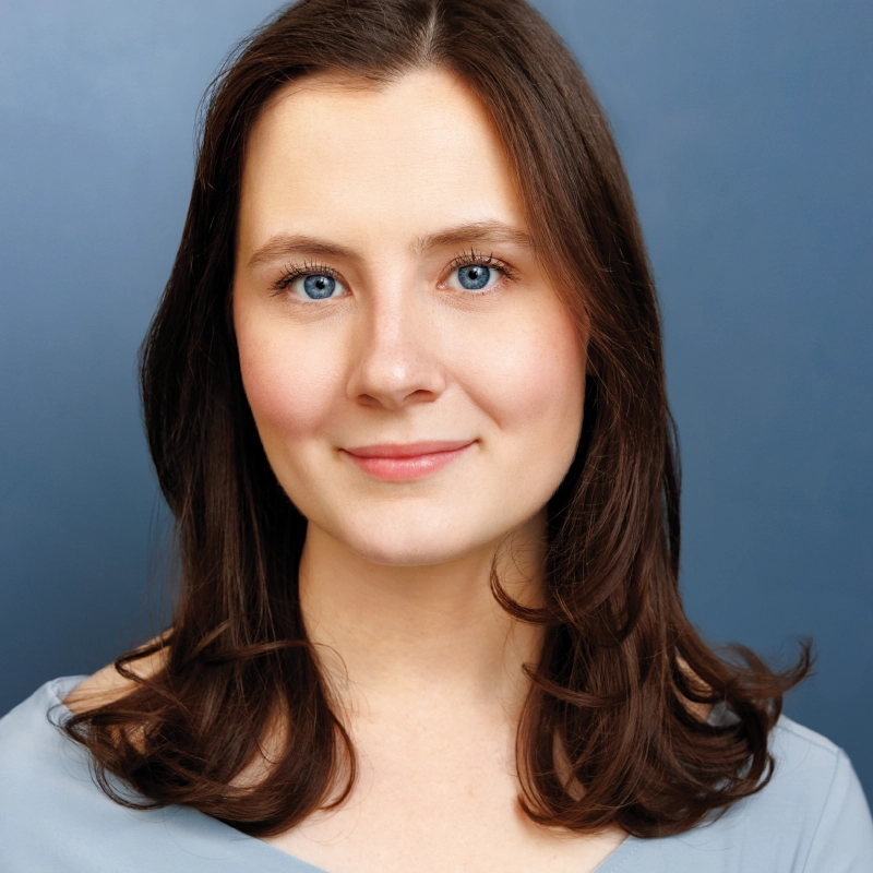 Katie Krasovec, a young woman with fair skin, blue eyes, and straight brown hair, smiles gently. She is wearing a light blue top against a solid blue background. The image features soft lighting and a professional, polished look.
