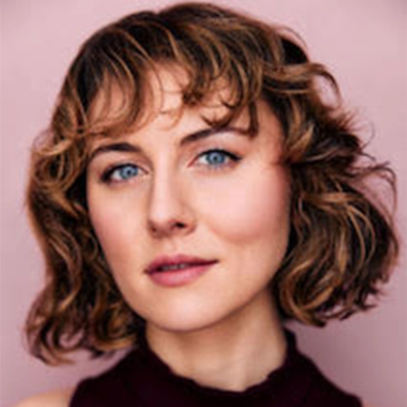 A woman with wavy, light brown, chin-length hair and blue eyes looks at the camera. Styled by Travis Flynt, she has fair skin, subtle makeup, and wears a sleeveless dark top against a soft pink background.