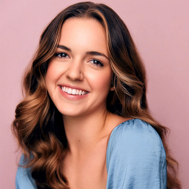 Katie Yeomans, a young woman with wavy brown hair and light highlights, smiles warmly. She wears a light blue top and has clear skin and blue eyes. The soft pink background gives the image a cheerful and friendly feel.