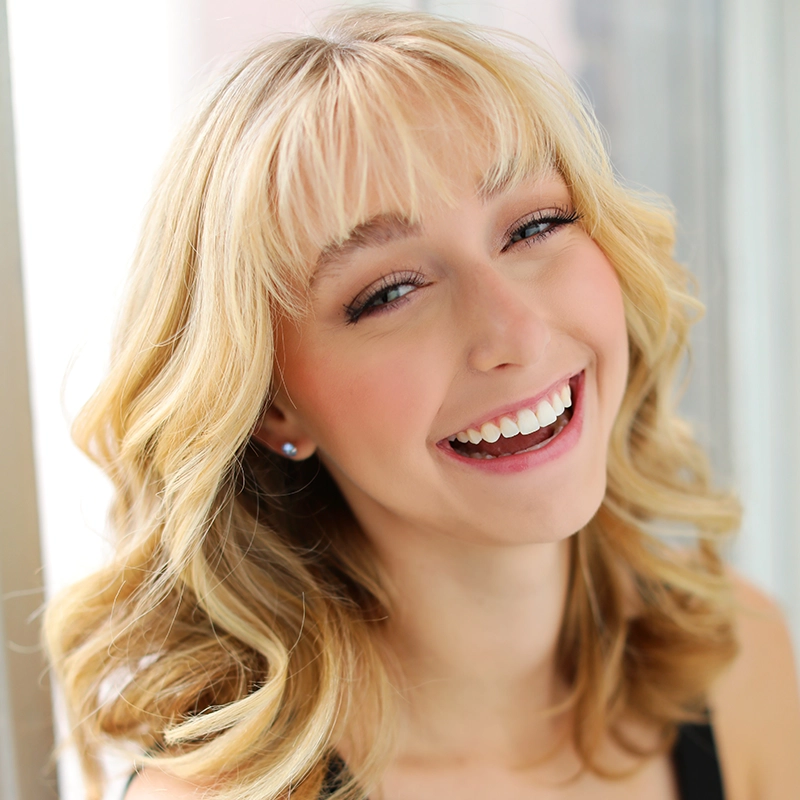 A young woman with wavy blonde hair and bangs smiles brightly, showing her teeth. She has fair skin, light makeup, and wears a sleeveless top. The softly blurred background suggests natural light—meet Kenna Wilson by the window.