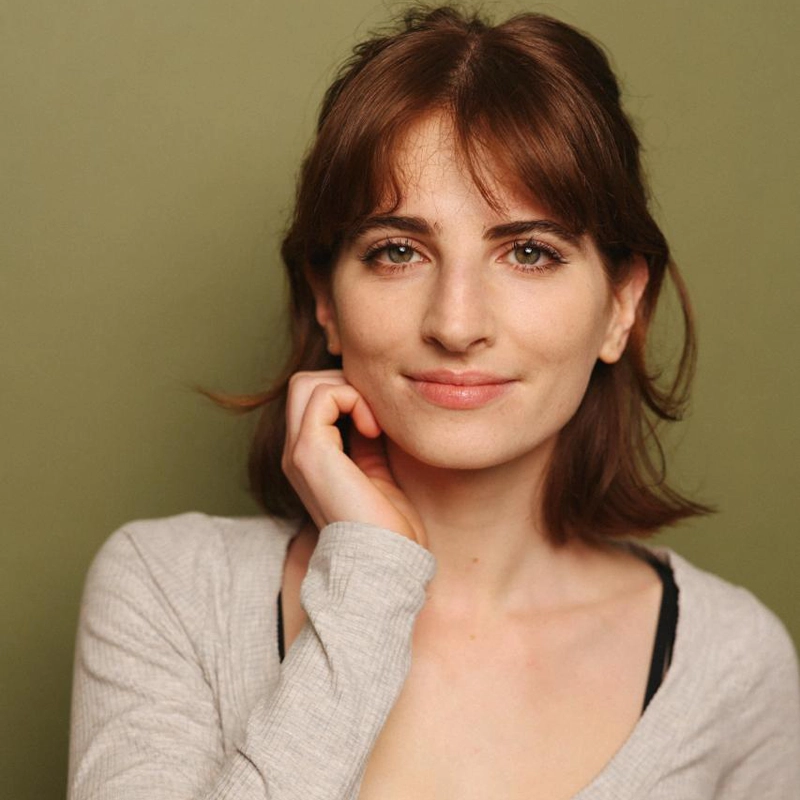 Rebecca Kent, a young woman with fair skin and brown, shoulder-length hair, stands against a plain olive green background. She wears a light gray top and looks at the camera with a gentle smile, resting her left hand on her face.