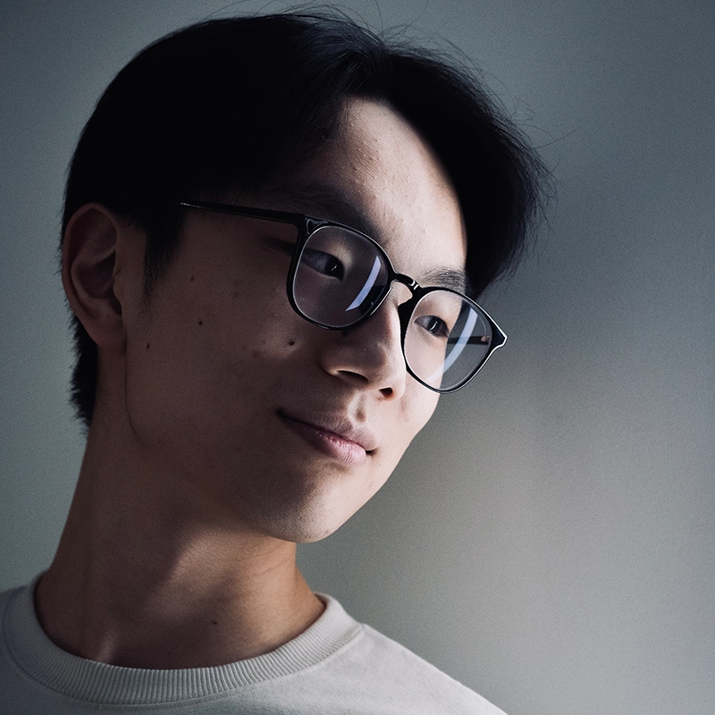A young person with short black hair and glasses looks to the side, softly smiling. They wear a light-colored shirt, and the background is plain and softly lit with a gradient from light to dark gray.