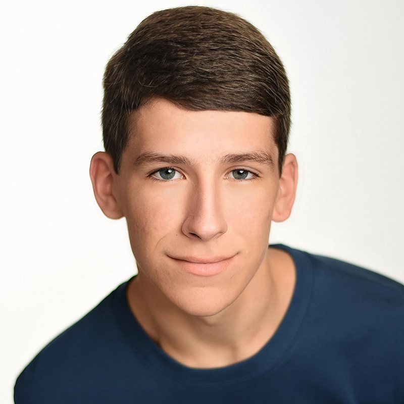 Lyla Karekinian, a young person with short brown hair and light skin, looks directly at the camera, smiling slightly. They wear a dark blue shirt against a plain, bright background with soft, even lighting.