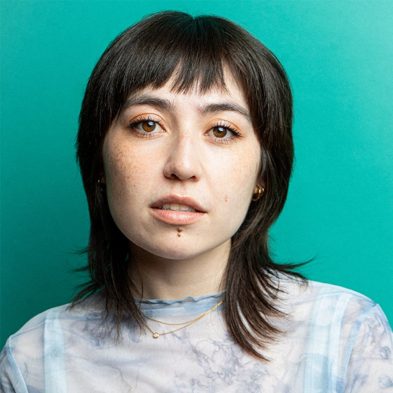 A young woman with straight, dark brown hair and bangs faces the camera against a teal background, styled in a look inspired by Aidan Michael Kelly. She has freckles, brown eyes, gold jewelry, and a facial piercing below her lip.