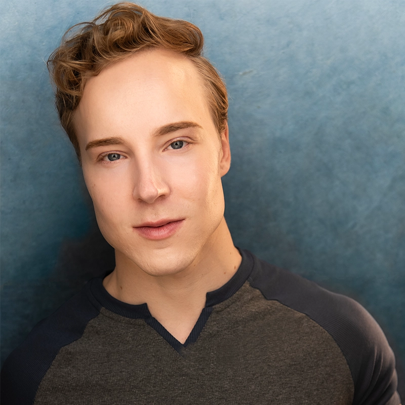 A young man with light skin, short wavy blond hair, and blue eyes is facing the camera, slightly smiling. He wears a gray shirt with dark raglan sleeves. The soft blue gradient background gives this Thompson portrait a calm and professional feel.