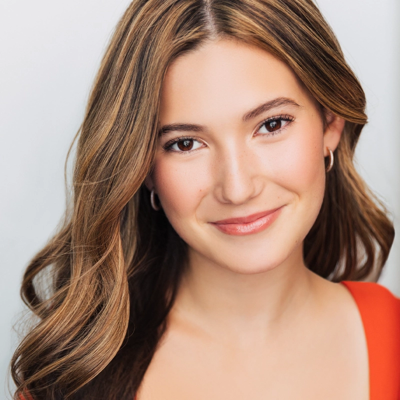Caitlyn Kops, a young woman with long, wavy light brown hair and fair skin, smiles gently. She wears a red top and small hoop earrings. Her natural makeup and the softly lit background highlight her face.