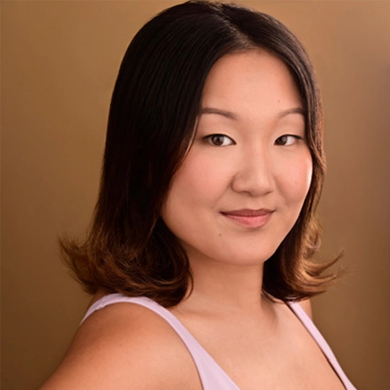 A young woman with medium-length, straight dark hair and fair skin, wearing a light pink sleeveless top, stands against a plain, warm brown background, looking confidently at the camera with a slight smile—reminiscent of Kourtnie Dwornikowski.