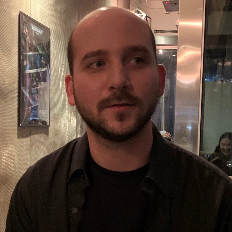 A man with a bald head and trimmed beard sits indoors, wearing a black shirt. The background includes a framed poster on a textured wall and a reflective panel. Dim lighting creates a cozy, relaxed atmosphere. Henryk Kress appears faintly in the background.