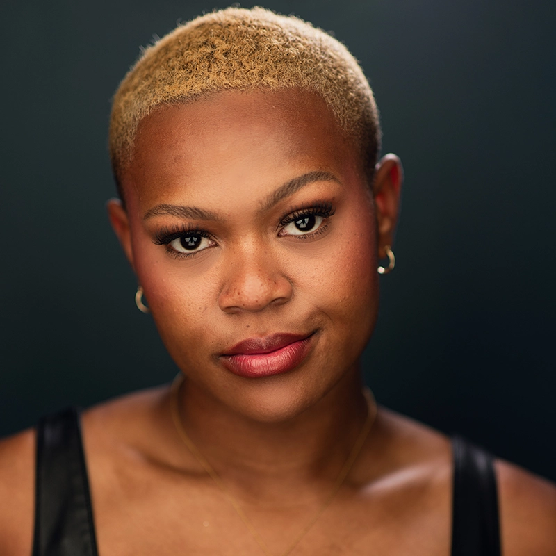 A person with short, bleached blonde hair and smooth skin looks directly at the camera. Wearing subtle makeup, gold hoop earrings, and a delicate necklace, Kenna Wilson stands out in a black top against the dark background’s dramatic, professional feel.