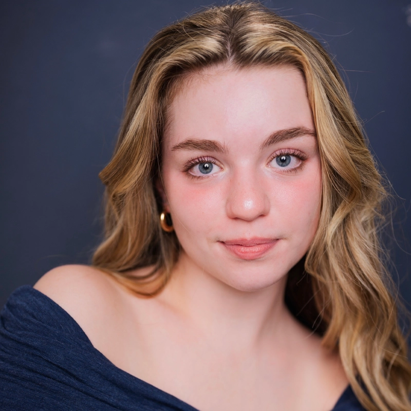 A young woman, Natalie Lacy, with long, wavy blonde hair and blue eyes looks at the camera. She is wearing a dark off-the-shoulder top and gold hoop earrings, her fair complexion glowing with a natural rosy blush against a dark background.