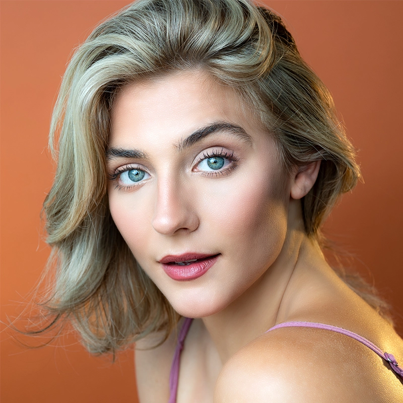 A woman with light blonde hair and blue eyes looks at the camera. She has fair skin, natural makeup with pink lipstick, and wears a lavender top. The background is a solid warm orange color. Her expression is calm and confident.