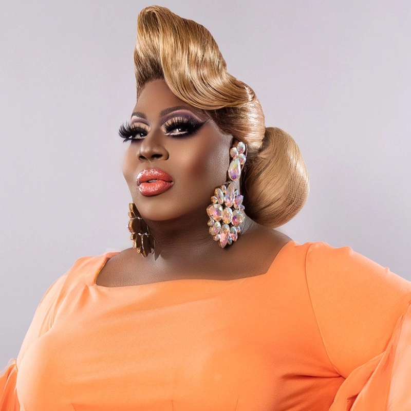 A person poses confidently, channeling Latrice Royale in a peach-colored outfit with dramatic, glossy makeup and thick false lashes. Their hair is styled in a sculpted, vintage updo with large, sparkling floral earrings against a plain background.