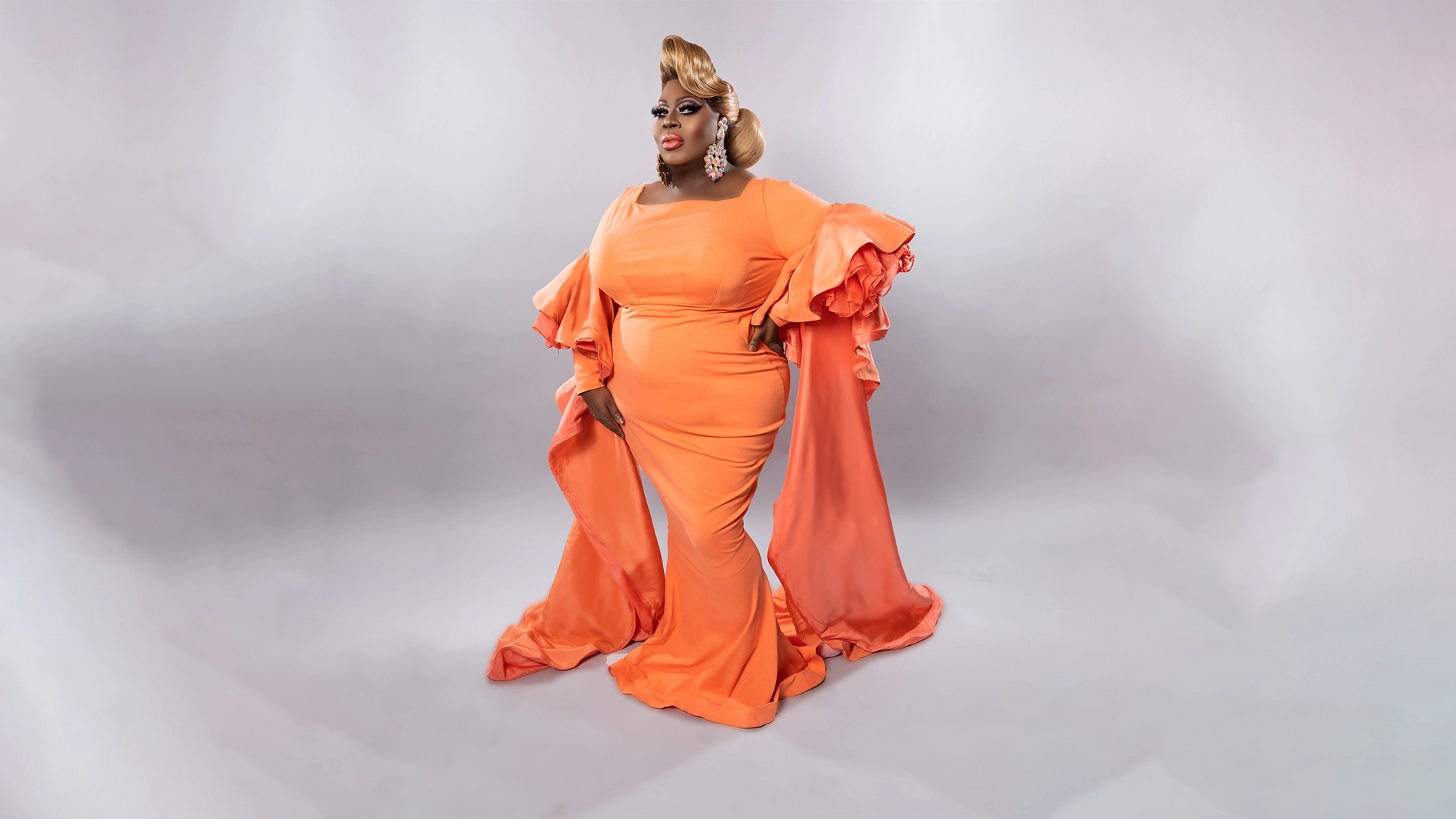 A person poses confidently in a floor-length, bright orange gown with dramatic ruffled sleeves, channeling Latrice Royale. They have styled blonde hair, bold makeup, and large sparkling earrings in a softly lit white studio setting.