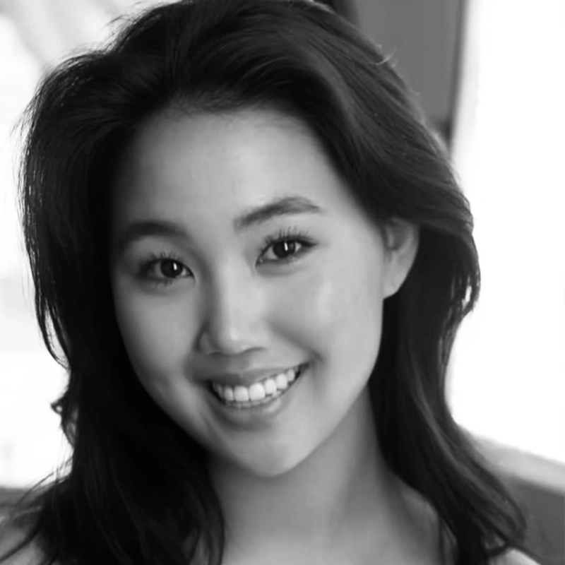 A young woman with long, dark hair smiles warmly at the camera. She has clear skin, expressive eyes, and is lit by soft, natural light, with a blurred background that enhances her friendly, approachable expression. The photo is in black and white.