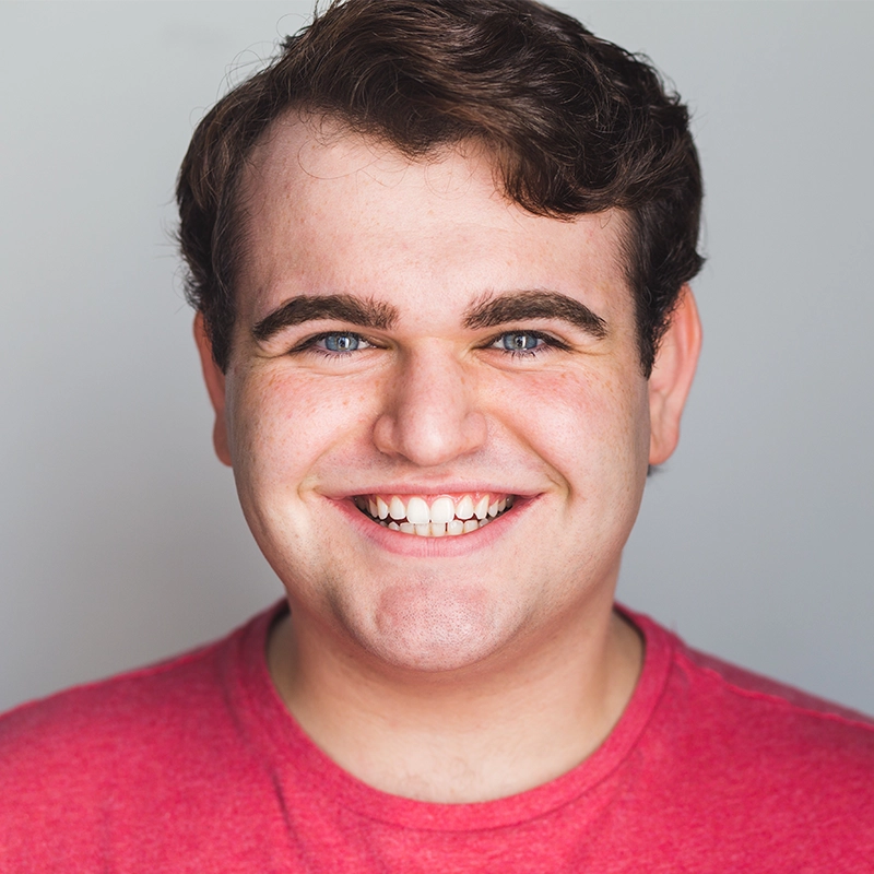A young man with short brown hair and blue eyes smiles broadly, showing his teeth. Wearing a red T-shirt, he stands against a plain light gray background. With a fair complexion, he appears happy and cheerful—captured perfectly by Kenna Wilson.