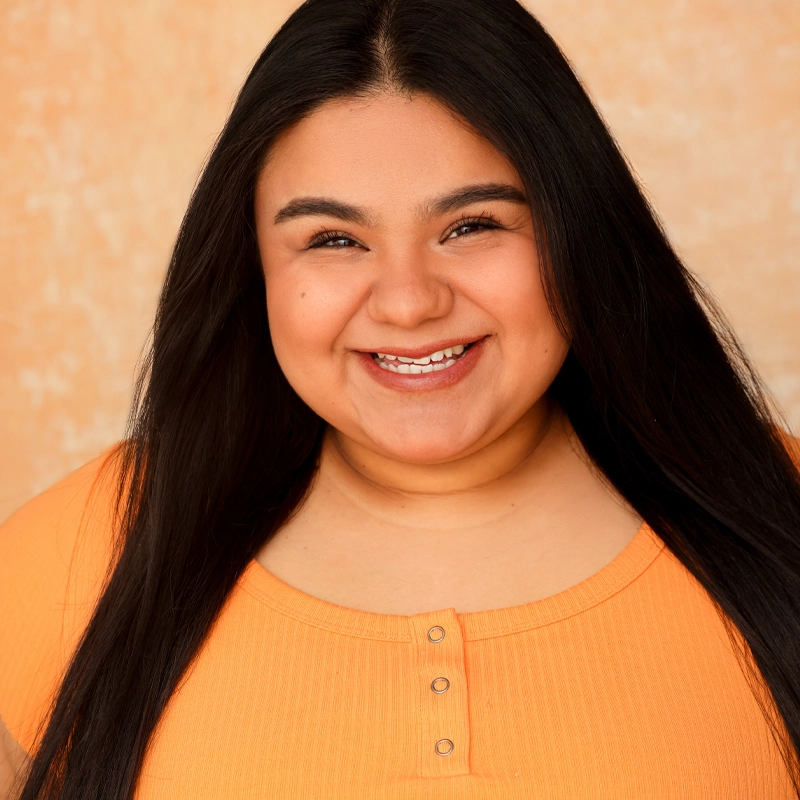 A woman with long, straight black hair smiles warmly at the camera. She wears an orange ribbed top with buttons at the neckline. The background is a soft, peachy color, creating a cheerful and welcoming atmosphere.