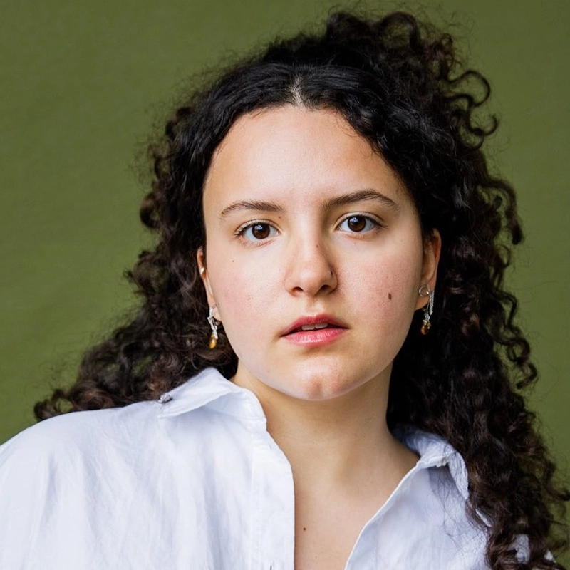 A young woman with curly dark hair, wearing a white button-up shirt and small earrings, looks directly at the camera with a neutral expression. The background is a plain olive green, highlighting Lily Gilan James’s calm presence.