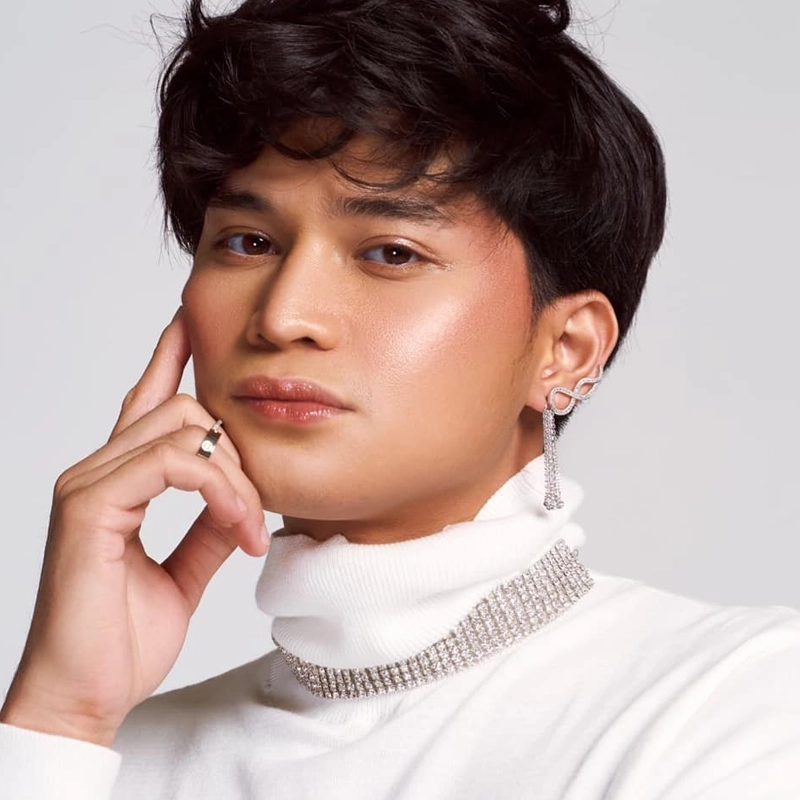 Adrian Lindayag, with short, wavy dark hair, wears a white turtleneck top, a sparkly choker, and a dangling earring. Their skin glows with soft makeup—rosy cheeks and glossy lips—as they gently touch their face and look confidently at the camera.