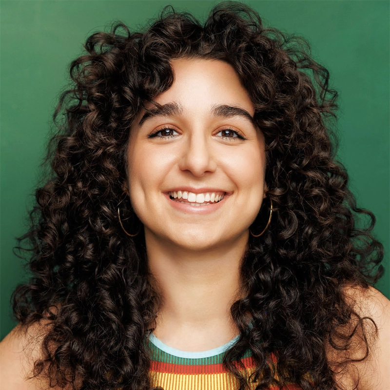 A young person with long, dark, curly hair smiles at the camera. Sasha Lippis wears gold hoop earrings and a colorful sleeveless top. The background is solid green, creating a bright and cheerful portrait vibe.