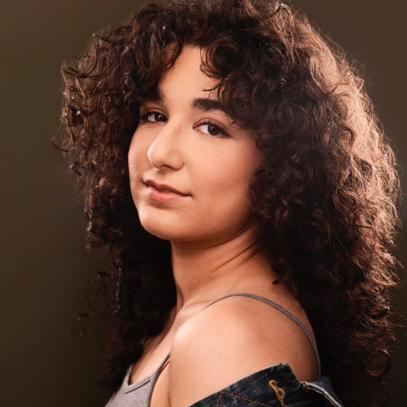 Sasha Lippis, a young woman with curly dark brown hair, poses against a dark background. She wears a gray spaghetti strap top with a jean jacket draped off her shoulder, gazing over her shoulder at the camera with gentle confidence.