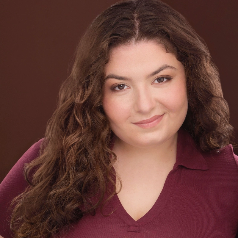 A woman with long, wavy brown hair and light skin smiles softly. She wears a maroon, collared top and natural makeup. The background is a solid, medium brown color, and the lighting highlights her face evenly.