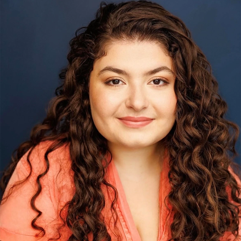 Woman with long, curly brown hair smiles softly at the camera. She wears a peach blouse and has natural makeup. The background is a solid dark blue, creating contrast with her hair and outfit. Her expression is calm and approachable.