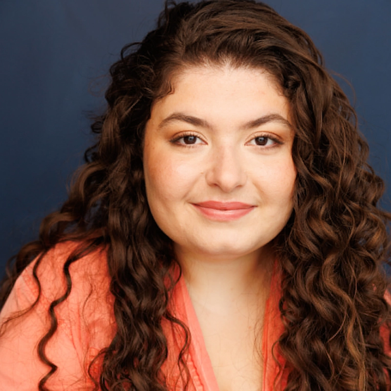 A woman with long, dark, curly hair and fair skin smiles softly. She is wearing a coral pink top and natural makeup, with a deep blue background behind her. Her expression is warm and friendly.