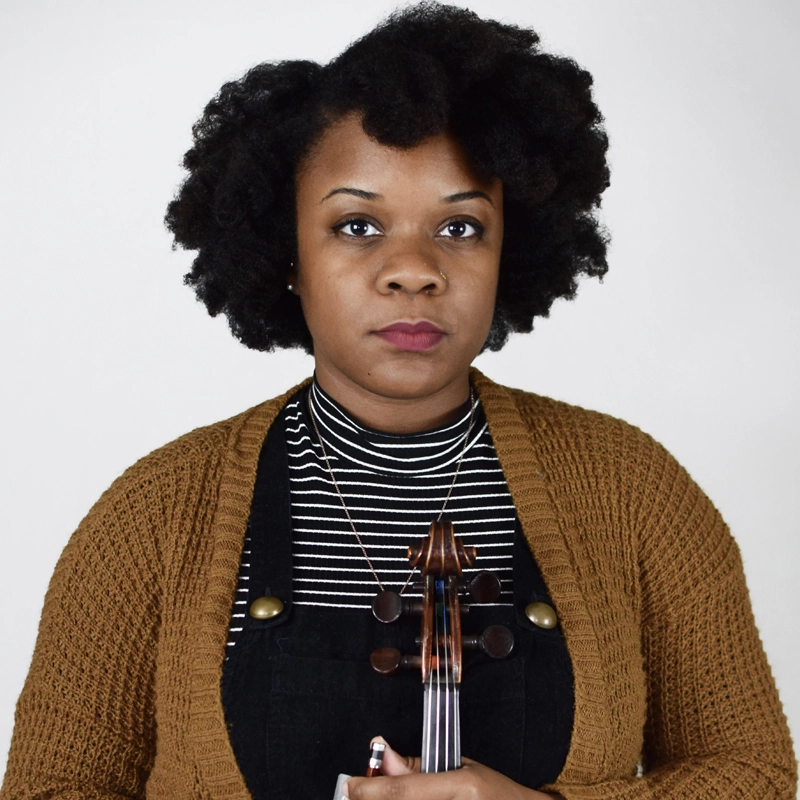 Leandria Lott, a woman with natural curly hair, wears a brown knit cardigan over a black and white striped shirt. She stands against a plain background, holding a violin upright in front of her chest and looking directly at the camera with a neutral expression.