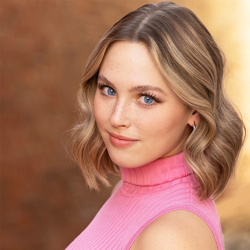 A young woman with wavy, shoulder-length blonde hair and blue eyes smiles softly. She wears a sleeveless, ribbed pink turtleneck. The background is blurred in warm brown tones, making her face the focal point of the image.