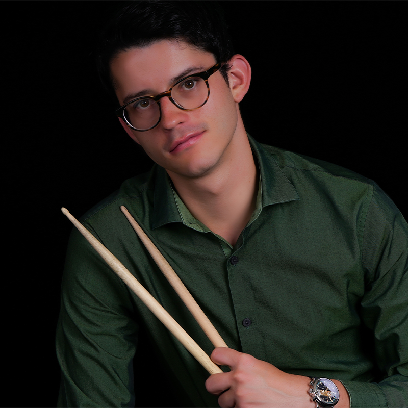 A young man wearing glasses and a green shirt holds two drumsticks. He is seated against a black background, looking at the camera with a neutral expression. He is also wearing a watch on his left wrist.