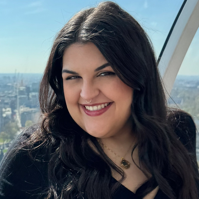 A woman with long, dark wavy hair smiles at the camera. She wears a black top, a gold necklace, and a nose ring. The background shows a cityscape and blue sky through a large window, suggesting she is in a high-rise or observation area.