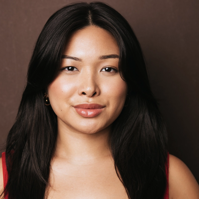 Miranda Macasero, with long, straight black hair and light makeup, smiles softly. She wears a red top and gold hoop earrings. Against a dark brown background, her calm and confident expression shines as she looks directly at the camera.