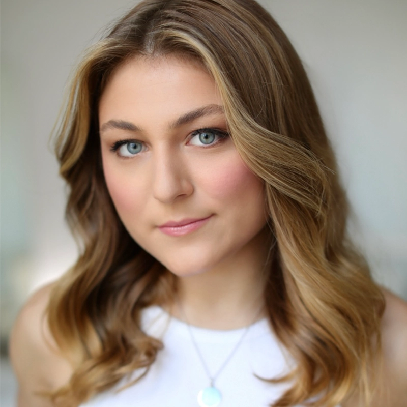 Kenna Wilson, a young woman with wavy, light brown hair and blue eyes, looks at the camera with a soft smile. She wears a white sleeveless top and a silver necklace with a round pendant. The softly blurred background draws focus to her face.