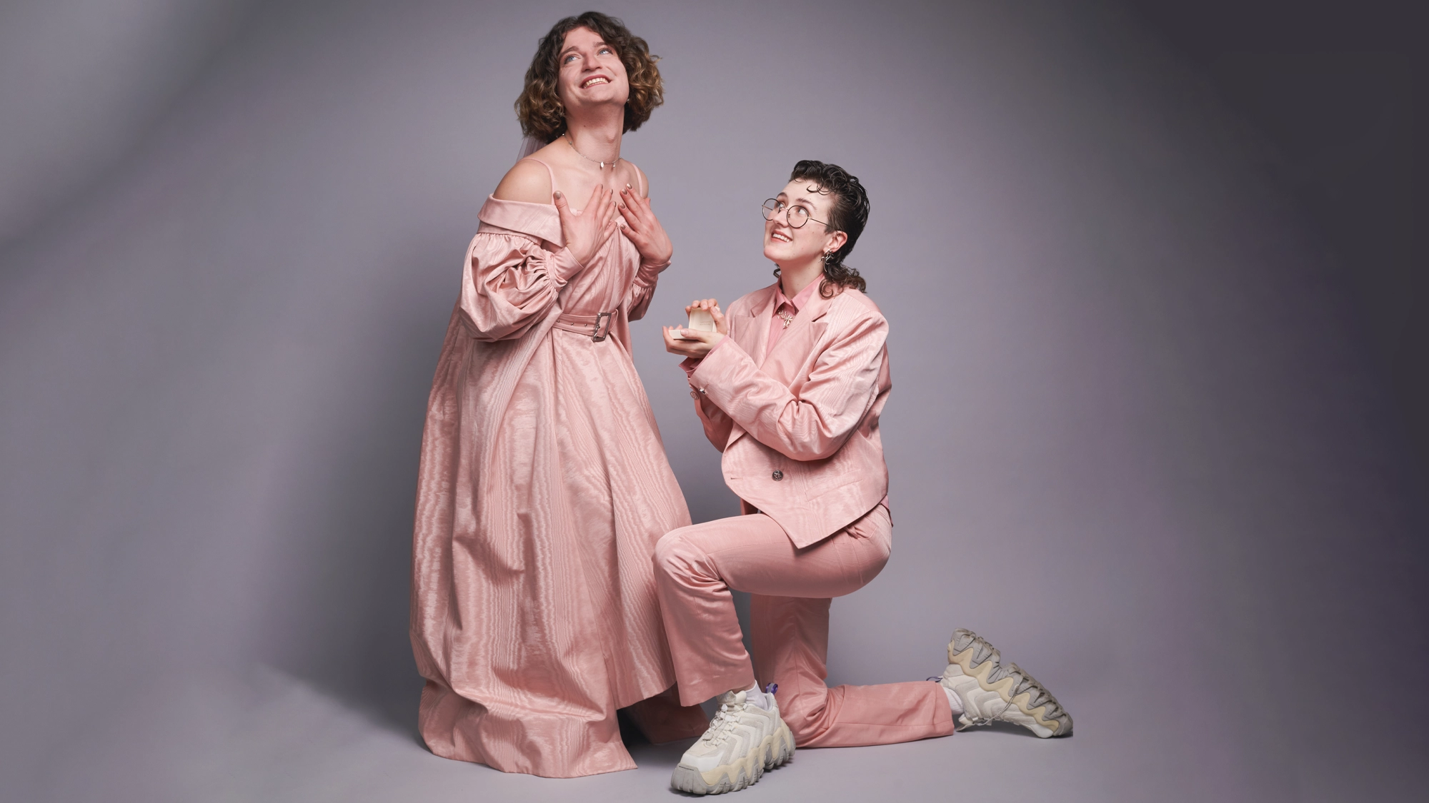 Two people pose against a gray backdrop in a playful engagement scene. Dressed in pink, they radiate joy—like Marlow and Moss from SIX, sharing a lighthearted moment that could answer the question: Why Am I So Single?.
