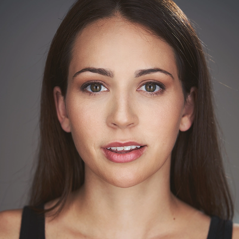 A young woman with long brown hair, resembling Abby Wolanin, looks directly at the camera. She has brown eyes, natural makeup, and wears a black top. The simple dark background keeps the focus on her neutral, expressive face.