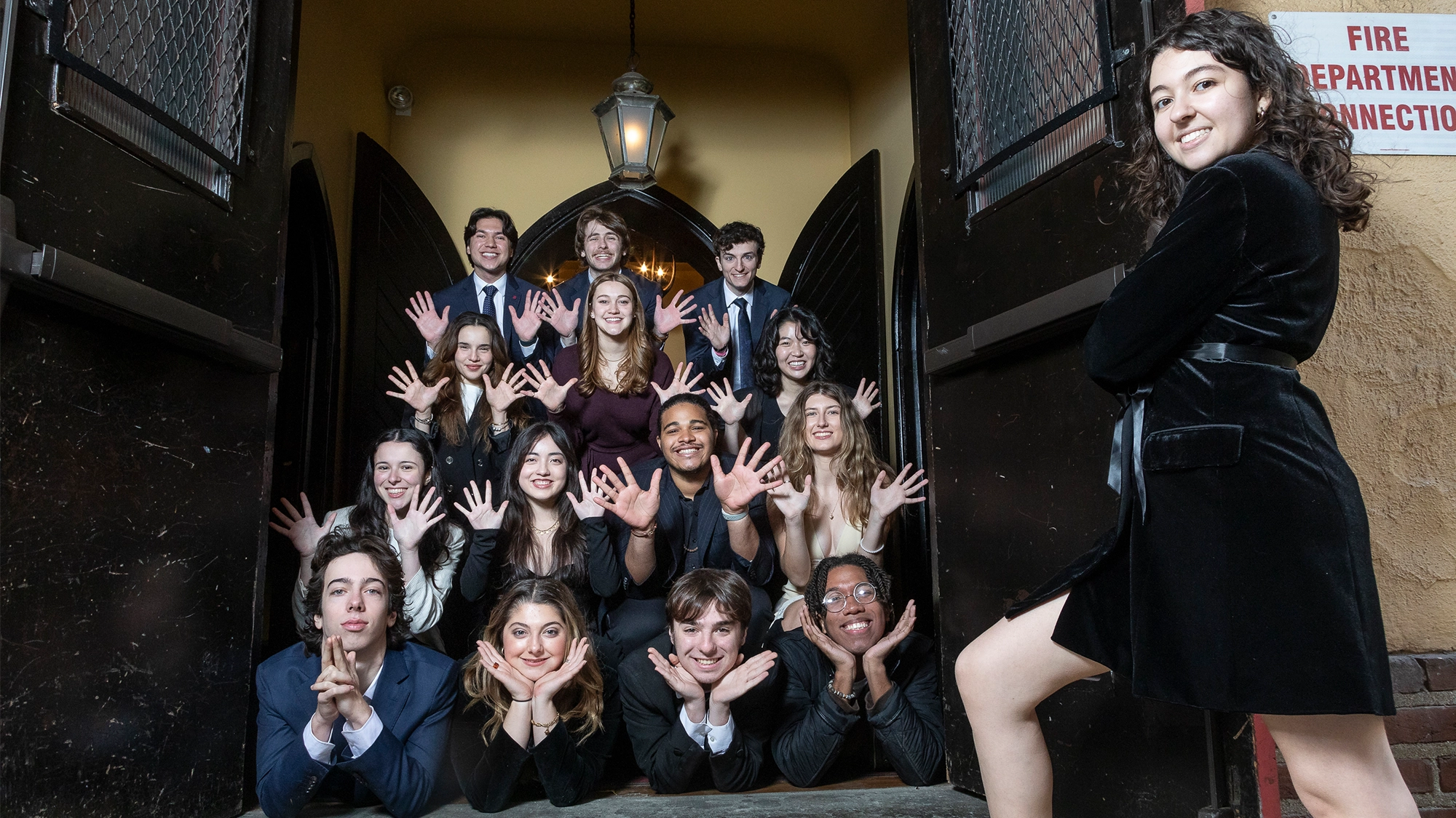 A group of 16 young adults dressed formally pose on steps outside double doors, many crouching with hands splayed near their faces in a playful Wiggin’ Out spirit. One woman in a black dress stands at the right, smiling confidently beneath a hanging lantern.