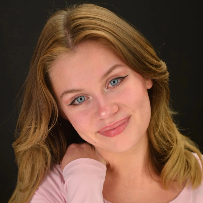 A young woman, Lizzie Matlock, with light brown hair and blue eyes smiles softly. She wears light pink makeup and a pale pink top, with one hand touching her neck. The black background makes her face and expression the focal point.