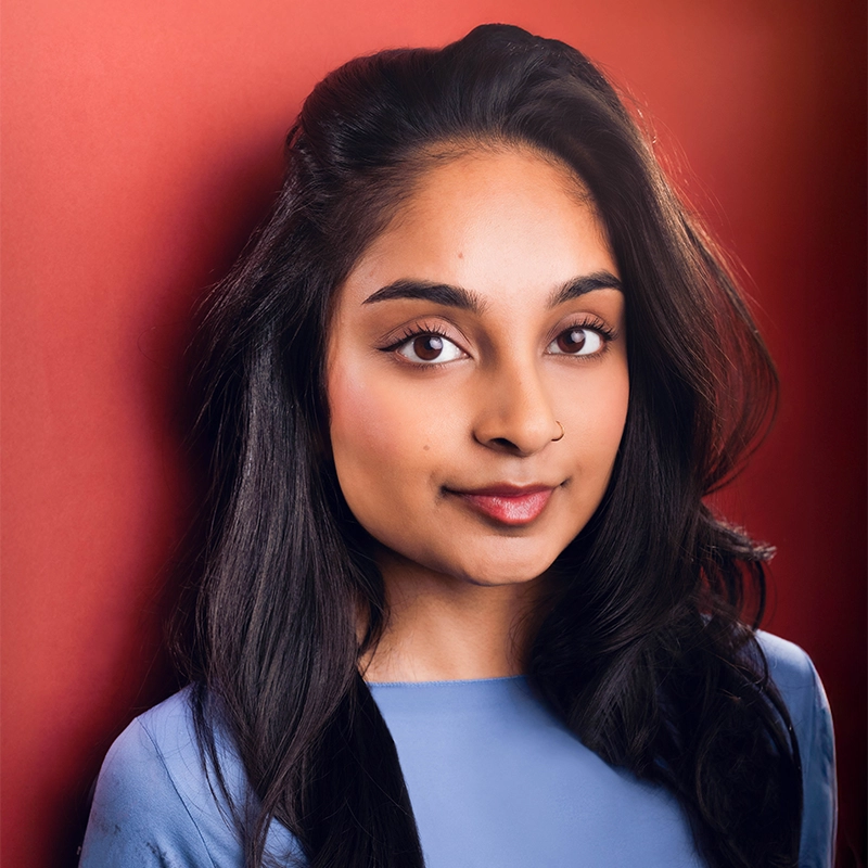 Maya Muthuramalingam, a young woman with long, dark hair and medium skin tone, poses against a deep red background in a light blue top. She gazes confidently at the camera, wearing natural makeup and a small nose ring.
