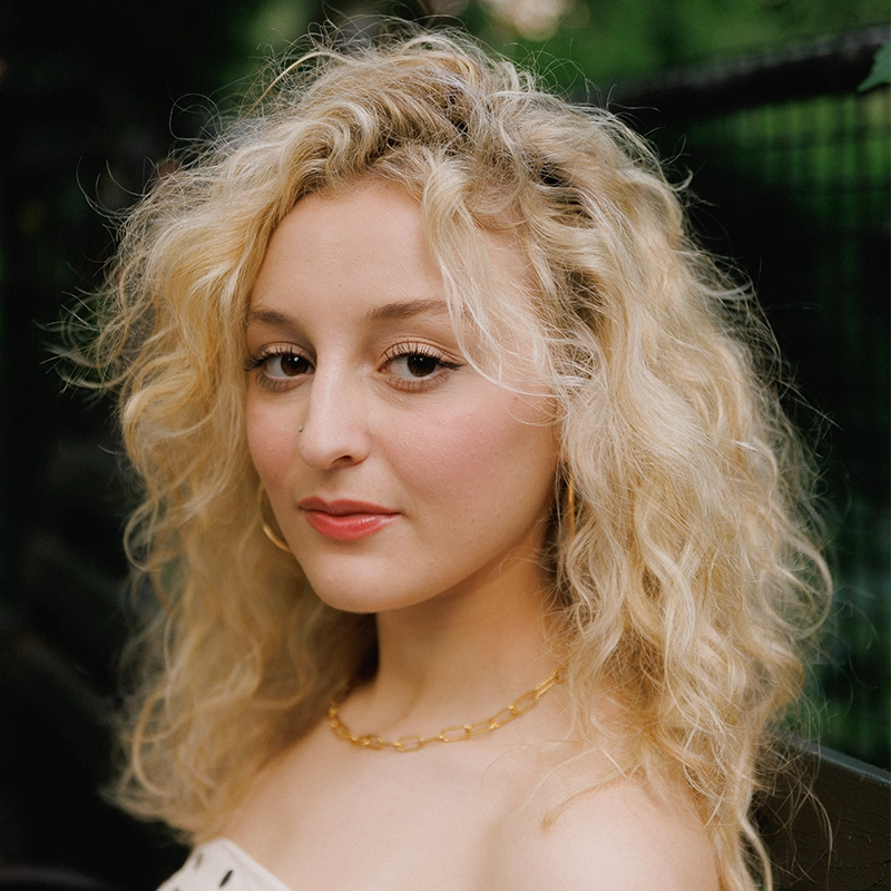 A woman with pale skin and long, curly blonde hair looks at the camera. She wears a gold chain necklace and hoop earrings. Her makeup includes subtle eyeliner and pink lipstick. The background is blurred greenery and a black fence, creating an elegant setting that highlights Ireyssa Cardona’s striking presence.